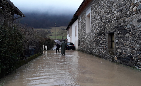 Rue du village inondée, avec deux passants équipés de bottes et d'un parapluie